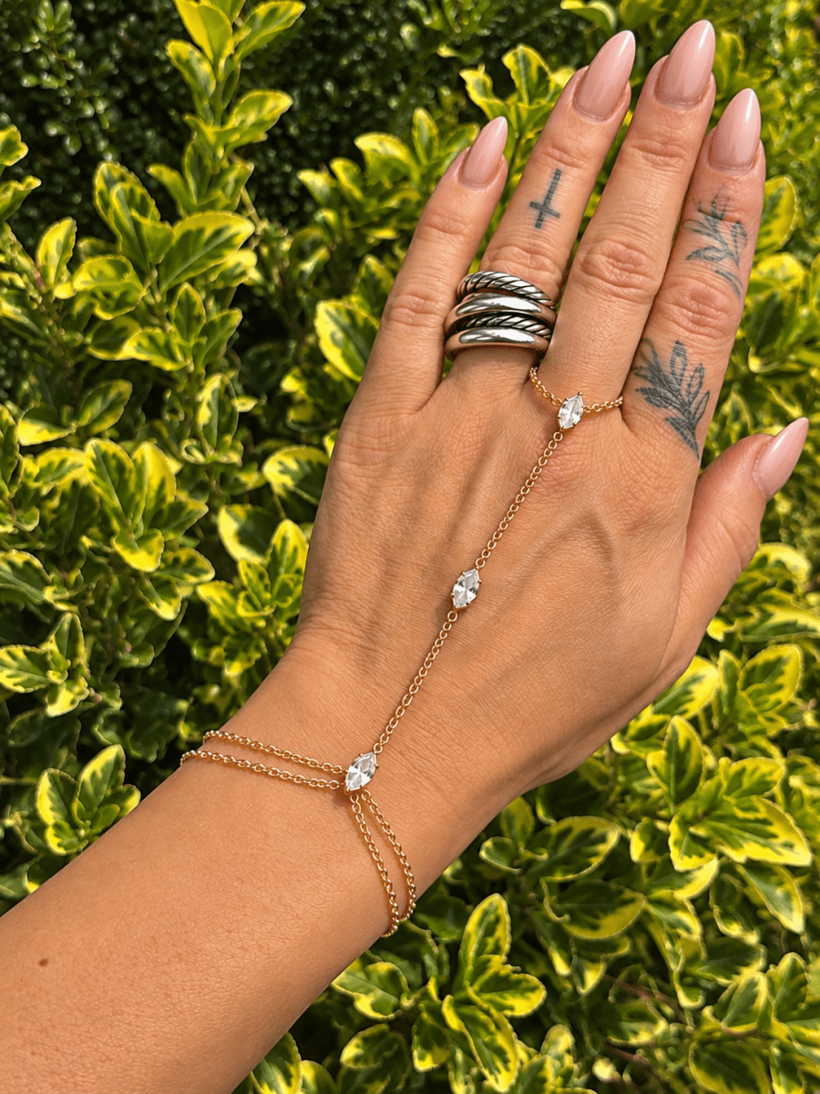Crystal Hand Chain Bracelet | Gold or Silver Finger Ring Bracelet Wristlet | bracelet | A gold chain hand bracelet with three clear crystal drops is shown on a person's hand, which is resting against a background of green and yellow variegated foliage.
