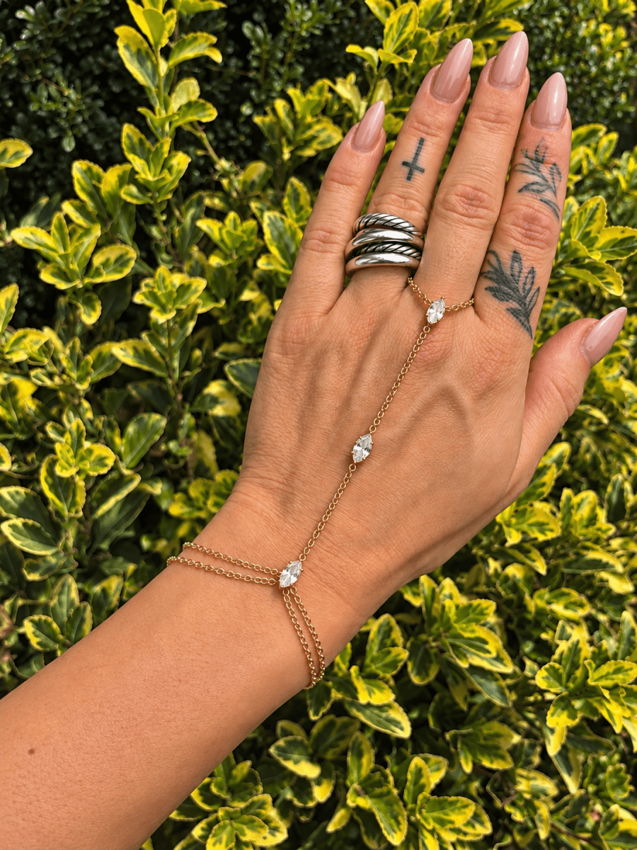 Crystal Hand Chain Bracelet | Gold or Silver Finger Ring Bracelet Wristlet | bracelet | A gold chain hand bracelet with three clear gemstone accents is shown on a person's hand, which is decorated with silver rings, against a background of green and yellow variegated garden foliage.