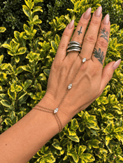 Crystal Hand Chain Bracelet | Gold or Silver Finger Ring Bracelet Wristlet | bracelet | A gold chain hand bracelet with three clear gemstone accents is shown on a person's hand, which is decorated with silver rings, against a background of green and yellow variegated garden foliage.