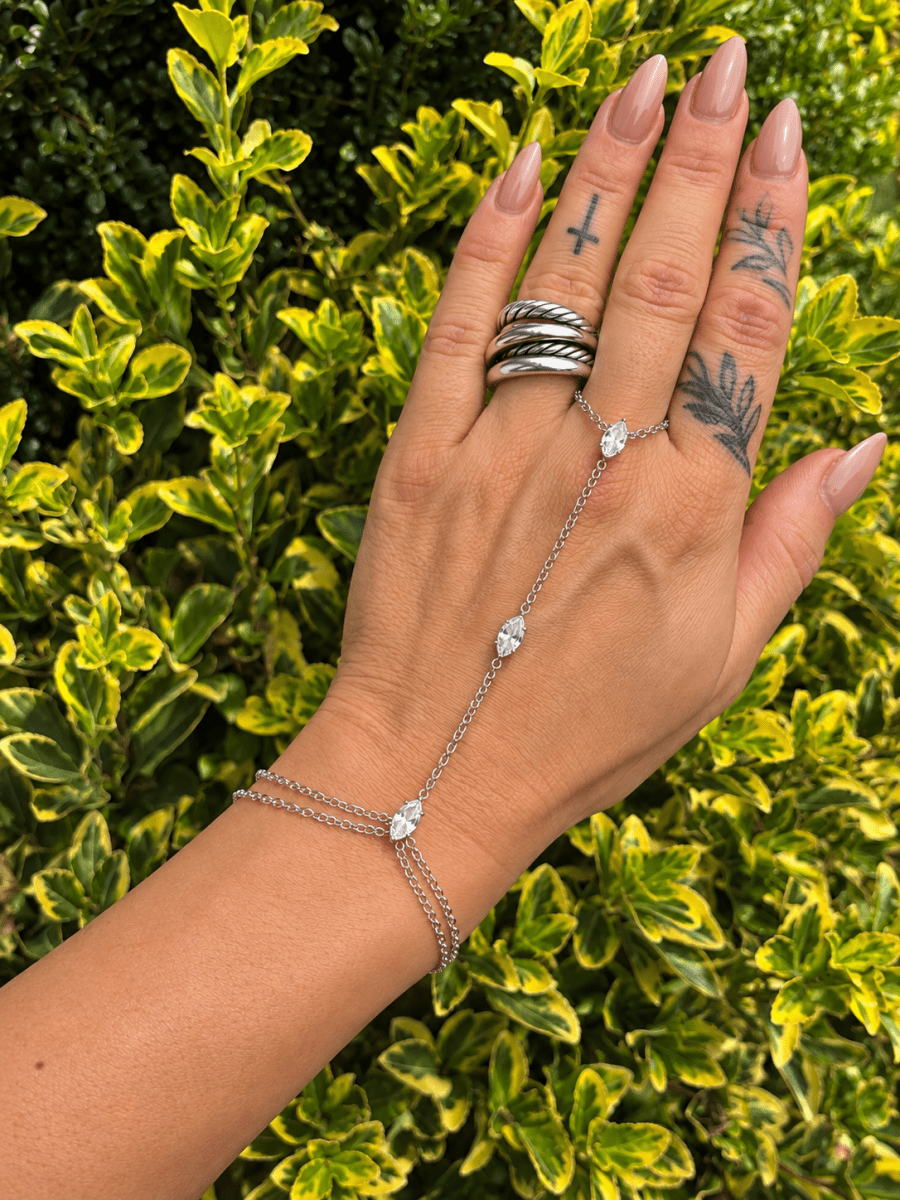 Crystal Hand Chain Bracelet | Gold or Silver Finger Ring Bracelet Wristlet | bracelet | A silver chain hand bracelet with three clear crystal drops is shown on a person's wrist, with their hand resting against a background of green and yellow variegated holly leaves. The hand also features silver rings on the fingers.