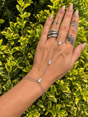 Crystal Hand Chain Bracelet | Gold or Silver Finger Ring Bracelet Wristlet | bracelet | A silver chain hand bracelet with three clear crystal drops is shown on a person's wrist, with their hand resting against a background of green and yellow variegated holly leaves. The hand also features silver rings on the fingers.