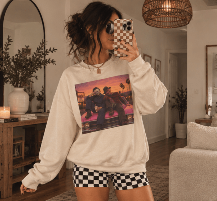 A woman takes a mirror selfie in a cozy living room, wearing a cream-colored sweatshirt with a colorful graphic print of two men sitting on a car, paired with black and white checkered shorts.